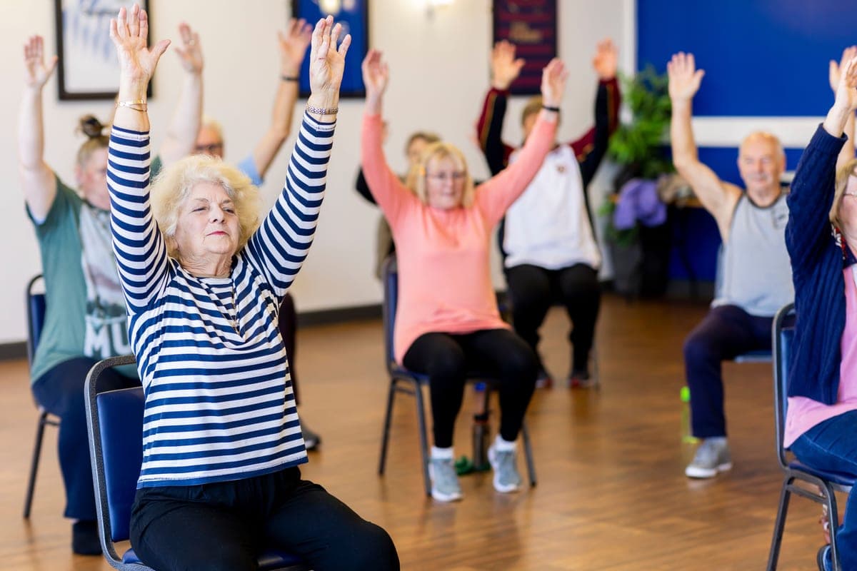 An older woman doing a standing stretch at home with a chair within reach